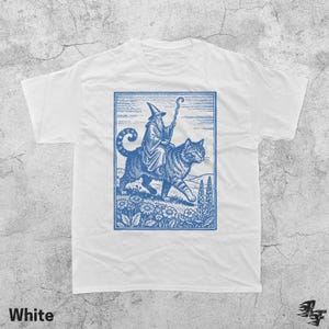 May include: White t-shirt with a blue graphic of a wizard riding a cat, holding a staff. The graphic is in a rectangular frame with flowers and a landscape background. The word "White" is printed at the bottom.