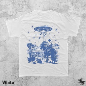May include: White t-shirt with a blue graphic of a cowboy on horseback being abducted by a UFO. The design includes a desert landscape, stars, and the word "White" at the bottom.