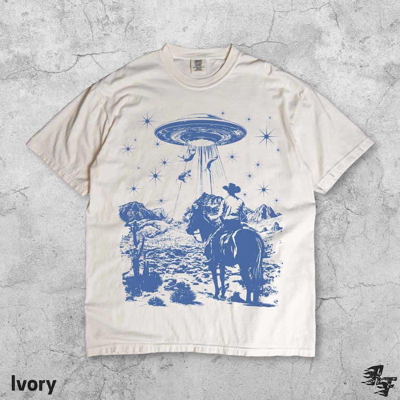 May include: Ivory-colored t-shirt with a blue graphic of a cowboy on a horse being abducted by a UFO. The design includes a desert landscape, stars, and the word "Ivory" at the bottom.