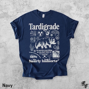 Puede incluir: Una camiseta azul marino con un estampado gr&aacute;fico blanco de un tard&iacute;grado rodeado de iconos cient&iacute;ficos y textos que incluyen "Tardigrade", "we survive everything so you don't have to", "Tiny, Tough & Nearly Immortal" y "outlived the dinosaurs".