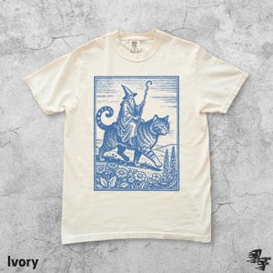 May include: Ivory-colored t-shirt featuring a blue illustration of a wizard riding a large cat through a field of flowers. The wizard wears a tall hat and holds a staff. The word "Ivory" is printed at the bottom left of the shirt.