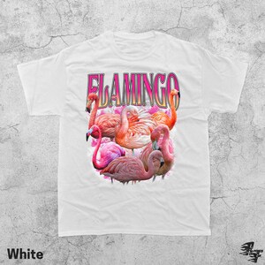 May include: White t-shirt featuring a vibrant graphic of several pink flamingos. The word "FLAMINGO" is printed in a pink and purple font above the birds. The word "White" is printed at the bottom of the shirt.