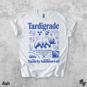 Puede incluir: Una camiseta gris claro con un estampado gr&aacute;fico azul de un tard&iacute;grado rodeado de iconos cient&iacute;ficos y el texto "Tardigrade, we survive everything so you don't have to, outlived the dinosaurs, simple math, Tiny, Nearly Immortal!"