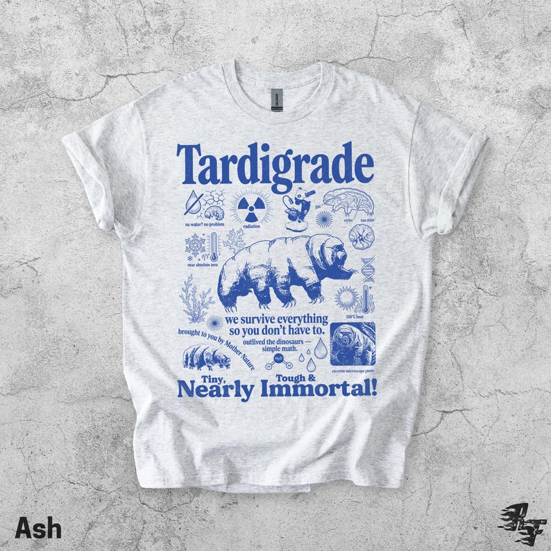 Puede incluir: Una camiseta gris claro con un estampado gr&aacute;fico azul de un tard&iacute;grado rodeado de iconos cient&iacute;ficos y el texto "Tardigrade, we survive everything so you don't have to, outlived the dinosaurs, simple math, Tiny, Nearly Immortal!"