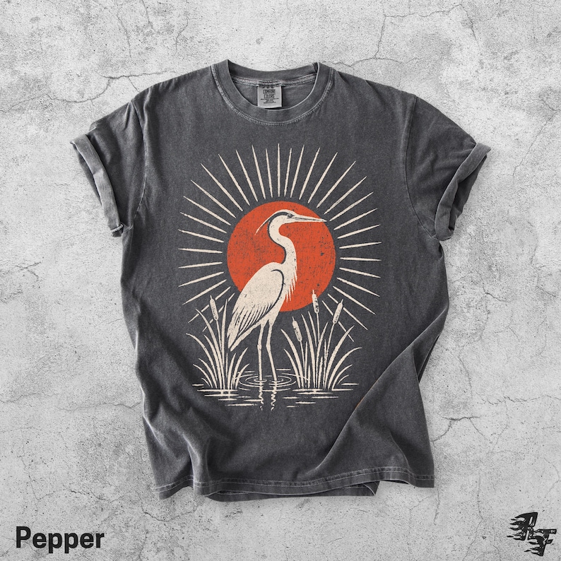 Pu&ograve; includere: Una t-shirt grigio antracite con una stampa grafica di un airone bianco in piedi tra i canneti davanti a un grande sole arancione con linee bianche radianti. La maglietta ha un girocollo, maniche corte e un'etichetta visibile sul colletto.