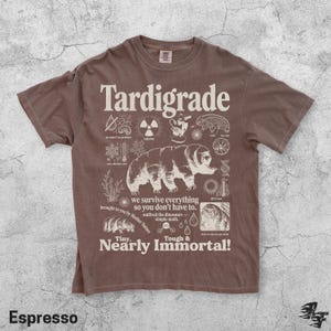 Puede incluir: Una camiseta marr&oacute;n de manga corta con un estampado gr&aacute;fico en color crema de un tard&iacute;grado. El dise&ntilde;o incluye iconos cient&iacute;ficos y el texto "Tardigrade", "we survive everything so you don't have to", "outlived the dinosaurs" y "Tiny, Nearly Immortal!"