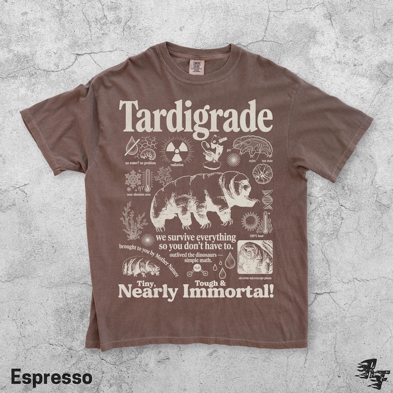 Puede incluir: Una camiseta marr&oacute;n de manga corta con un estampado gr&aacute;fico en color crema de un tard&iacute;grado. El dise&ntilde;o incluye iconos cient&iacute;ficos y el texto "Tardigrade", "we survive everything so you don't have to", "outlived the dinosaurs" y "Tiny, Nearly Immortal!"