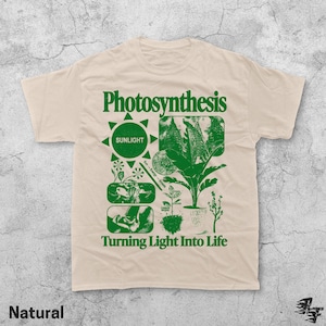Op de afbeelding: Natuurkleurig t-shirt met een groen grafisch ontwerp. Het ontwerp bevat het woord "Photosynthesis" boven een zon-icoon en botanische illustraties. De tekst "Turning Light Into Life" staat eronder.