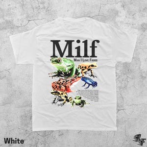 May include: White t-shirt with the text "Milf Man I Love Frogs" in black. The shirt features a colorful graphic of several frogs in various colors, including green, red, and blue. The shirt is made of a soft material.