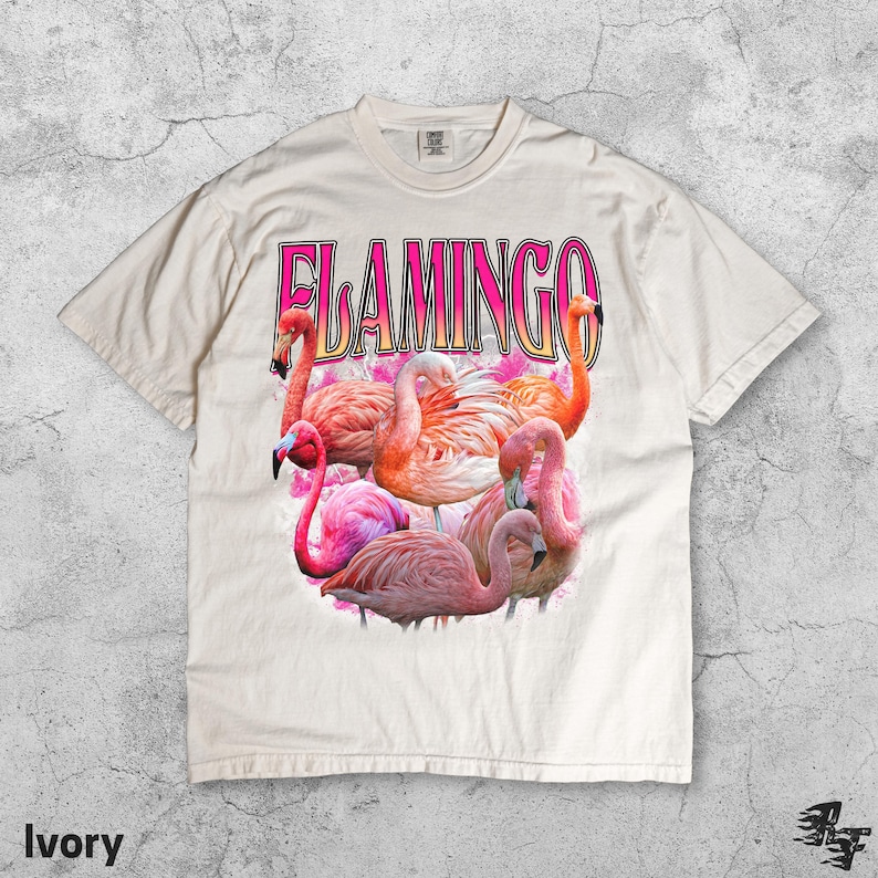 May include: Ivory-colored t-shirt featuring a graphic of several pink flamingos and the word "FLAMINGO" in pink letters. The shirt has a crew neck and short sleeves. The design is centered on the front of the shirt.