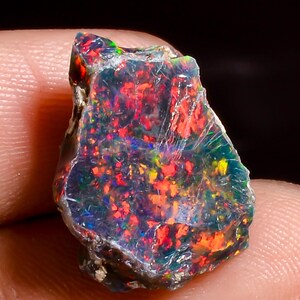 May include: A raw black opal gemstone featuring a dark base color with vibrant flashes of red, orange, and green play-of-color throughout its irregular, textured surface. The stone displays a natural, unpolished shape with visible crystalline inclusions.