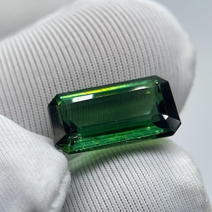 以下が含まれることがあります： 長方形のエメラルドグリーンの宝石で、ファセット加工されたエッジがあります。宝石は白い背景に配置され、その透明度と色を示しています。宝石の深い緑色は、その研磨された表面に反射する光によって強調されています。