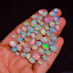 Puede incluir: Una colección de numerosos ópalos cabujón de forma ovalada y de lágrima con una base de color blanco lechoso y destellos vibrantes de fuego verde, naranja y azul. Estas piedras preciosas pulidas varían en tamaño y se muestran en una palma.
