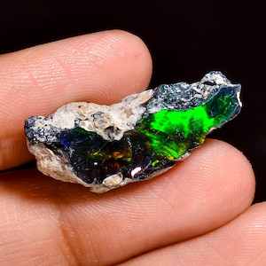 Peut inclure: Une opale noire brute et non polie présentant une base matricielle sombre et texturée, avec un jeu de couleurs irisé et vibrant composé d'éclats intenses de vert néon, de bleu et de touches de rouge sur sa surface cristalline fracturée.