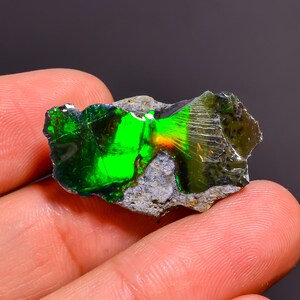 Peut inclure: Un spécimen d'opale brut et non poli présentant un jeu de couleurs vert néon vibrant sur sa surface. La pierre est incrustée dans une roche matricielle grise, rugueuse et texturée, affichant un contraste marqué entre le minéral vert brillant et réfléchissant et la base terreuse terne.