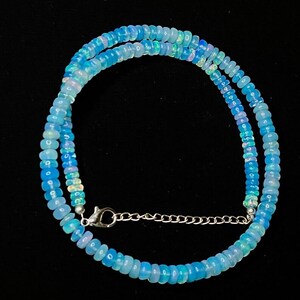 以下が含まれることがあります： 小さな丸い水色のオパールビーズで構成されたネックレス。ビーズはつながっており、シルバーカラーの留め金とチェーンで接続された二重のストランドを形成しています。背景は黒です。