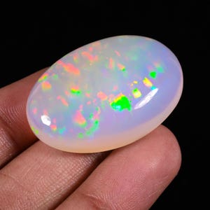 May include: A polished, oval-shaped opal gemstone. The opal displays a milky white base color with flashes of iridescent colors, including green, pink, and yellow. The gemstone is held in a hand against a black background.