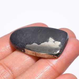 Pode incluir: Uma pedra preciosa polida, cinza escuro, com um brilho metálico e uma mancha de material prateado. A pedra tem uma forma retangular arredondada e é segurada em uma mão.