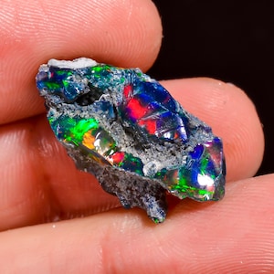 Peut inclure: Une pierre d'opale noire brute et irisée, avec des éclats vibrants de vert, de rouge et de bleu. L'opale est tenue dans une main, mettant en valeur sa texture naturelle non polie et ses jeux de couleurs. Un bijou naturel unique.