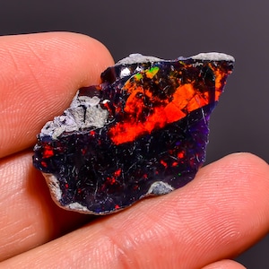 May include: A raw black opal gemstone featuring a dark, deep blue and black base color with vibrant, intense flashes of neon red and orange play-of-color across its surface, alongside patches of natural, rough grey stone matrix.
