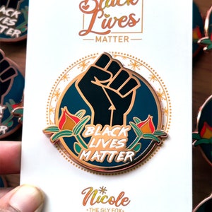 May include: A pin with a black fist in a blue circle, surrounded by gold accents and red and green floral designs. The words "Black Lives Matter" are displayed in gold lettering. The pin is on a white card with the text "Black Lives Matter" and "Nicole".