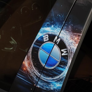 Op de afbeelding: Een autodeurstijl met een BMW-logo. Het logo is een blauw en wit cirkelvormig embleem met de letters "B", "M" en "W". De achtergrond heeft een blauw en oranje abstract ontwerp.