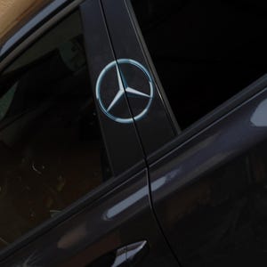 Op de afbeelding: Een close-up van een donkergrijze autodeur met een Mercedes-Benz logo. Het logo is een zilveren driepuntige ster in een zilveren cirkel. De deurklink is zichtbaar.