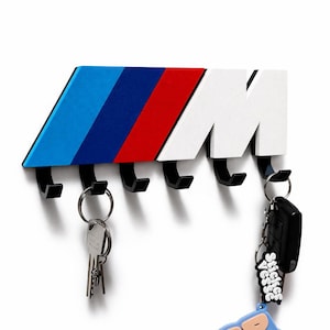 Könnte beinhalten: Ein Wand-Schlüsselhalter mit dem BMW M-Logo in Blau, Dunkelblau, Rot und Weiß. Er hat fünf schwarze Haken für Schlüssel und Schlüsselanhänger. Ein modernes und stilvolles Accessoire, um Schlüssel zu organisieren und griffbereit zu haben.