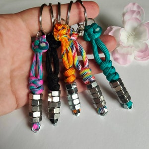 Peut inclure: Cinq porte-clés avec des accents d'écrous métalliques et des nœuds en paracorde colorés. Les porte-clés présentent une variété de couleurs, dont le turquoise, le noir, l'orange et un motif arc-en-ciel. Chaque porte-clés a un anneau argenté.