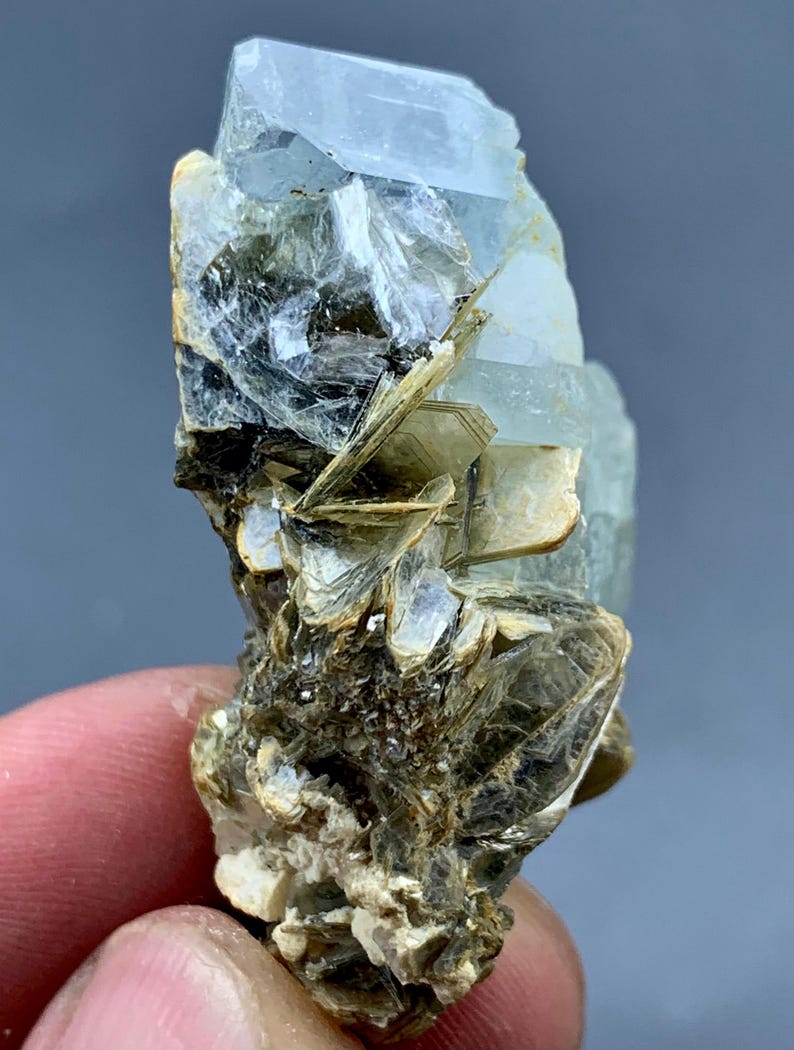 Peut inclure: Un amas de cristaux d'aigue-marine bruts, de couleur bleu p&acirc;le. Le cristal a une structure en couches avec une surface rugueuse et textur&eacute;e. La base de l'amas est un m&eacute;lange de min&eacute;raux bruns et beiges, cr&eacute;ant un aspect naturel et terreux.