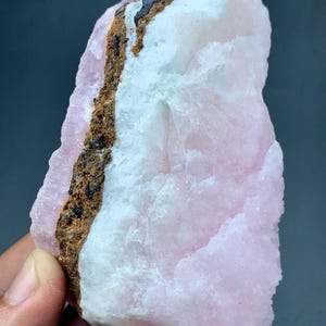 May include: A rough-cut, natural mineral specimen featuring a blend of pink, white, and brown hues. The stone has a textured surface with a prominent brown vein running through the center. The pink areas have a soft, opaque appearance.