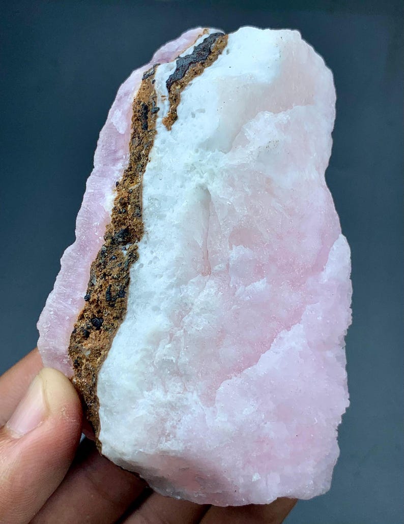 May include: A rough-cut, natural mineral specimen featuring a blend of pink, white, and brown hues. The stone has a textured surface with a prominent brown vein running through the center. The pink areas have a soft, opaque appearance.