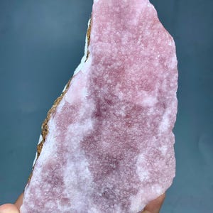 May include: A rough-cut, pink mineral specimen with a textured surface. The stone has areas of lighter pink and white, with a brown and white edge. The mineral is held in a hand against a blue-gray background.
