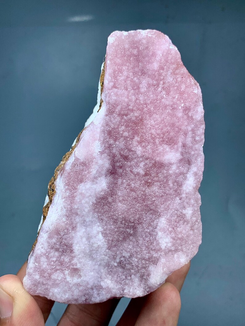 May include: A rough-cut, pink mineral specimen with a textured surface. The stone has areas of lighter pink and white, with a brown and white edge. The mineral is held in a hand against a blue-gray background.
