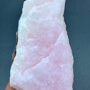 May include: A large, raw piece of pink calcite crystal. The stone has a rough, textured surface with varying shades of pink and white. The crystal is held in a hand against a dark background.