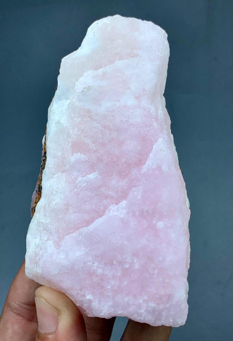 May include: A large, raw piece of pink calcite crystal. The stone has a rough, textured surface with varying shades of pink and white. The crystal is held in a hand against a dark background.