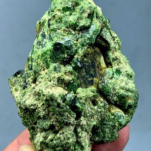 May include: A rough, natural mineral specimen with a vibrant green hue. The surface is textured and uneven, with crystalline formations and darker green inclusions. The overall shape is irregular, showcasing the raw beauty of the stone.