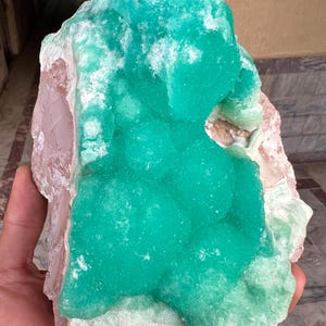 May include: A large, raw mineral specimen featuring a vibrant, teal-green crystalline formation. The mineral has a textured surface with areas of white and pinkish-brown hues. The overall shape is irregular and natural, showcasing the beauty of the raw stone.