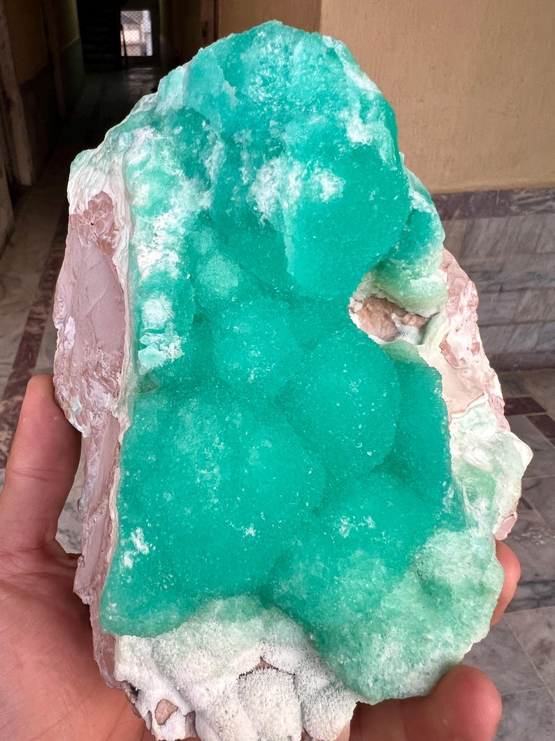May include: A large, raw mineral specimen featuring a vibrant, teal-green crystalline formation. The mineral has a textured surface with areas of white and pinkish-brown hues. The overall shape is irregular and natural, showcasing the beauty of the raw stone.
