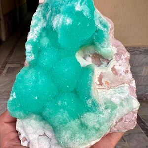 May include: A large, raw mineral specimen featuring a vibrant, crystalline, turquoise-green formation atop a matrix of pink and white stone. The mineral has a bubbly, textured appearance, and the overall piece is irregularly shaped.