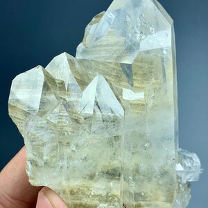 May include: A cluster of clear quartz crystals with a pale yellow hue. The crystals have sharp, geometric shapes and varying sizes, with a large, prominent central point. The overall structure is rough and natural.