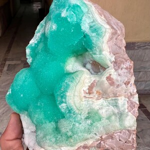 May include: A large, raw mineral specimen featuring a vibrant turquoise hue with areas of white and light brown. The crystalline structure has a layered appearance, with a rough, textured surface. The mineral is held in a hand.