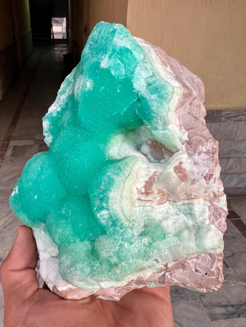 May include: A large, raw mineral specimen featuring a vibrant turquoise hue with areas of white and light brown. The crystalline structure has a layered appearance, with a rough, textured surface. The mineral is held in a hand.