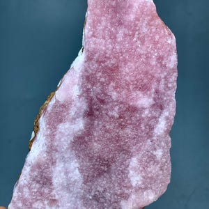 May include: A rough-cut, pink rhodochrosite mineral specimen. The stone has a textured surface with areas of lighter and darker pink hues. The edges show a mix of white and brown coloration. The overall shape is irregular and natural.
