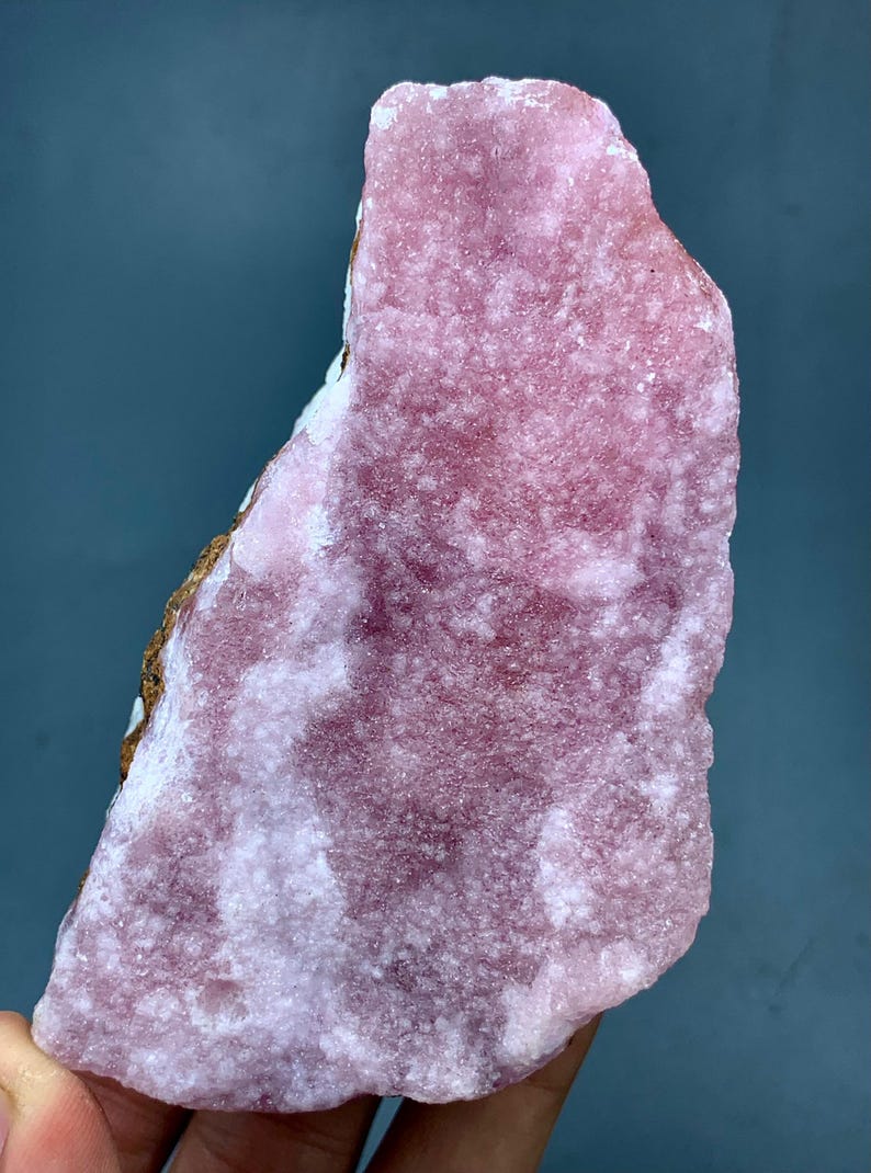 May include: A rough-cut, pink rhodochrosite mineral specimen. The stone has a textured surface with areas of lighter and darker pink hues. The edges show a mix of white and brown coloration. The overall shape is irregular and natural.
