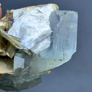 Peut inclure: Gros plan sur un cristal d'aigue-marine brut de couleur bleu p&acirc;le. Le cristal est partiellement int&eacute;gr&eacute; dans une matrice de min&eacute;raux blancs et beiges, cr&eacute;ant un aspect naturel et textur&eacute;. L'aigue-marine est translucide.