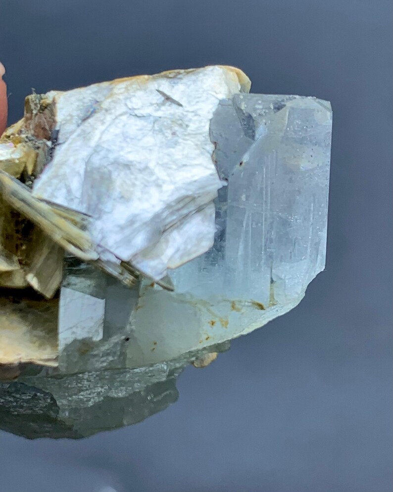Peut inclure: Gros plan sur un cristal d'aigue-marine brut de couleur bleu p&acirc;le. Le cristal est partiellement int&eacute;gr&eacute; dans une matrice de min&eacute;raux blancs et beiges, cr&eacute;ant un aspect naturel et textur&eacute;. L'aigue-marine est translucide.
