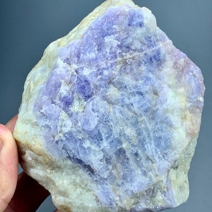 May include: A raw, natural gemstone with a blend of lavender and cream hues. The stone's surface displays a crystalline structure with variations in color intensity, ranging from pale lavender to deep purple. The rough texture and irregular shape highlight its natural origin.