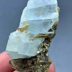 Peut inclure: Sp&eacute;cimen brut de cristal d'aigue-marine. Le cristal bleu p&acirc;le est partiellement transparent et repose sur une matrice de min&eacute;raux bruns et beiges. Le cristal a un aspect stratifi&eacute; avec des facettes visibles et une texture naturelle non polie.