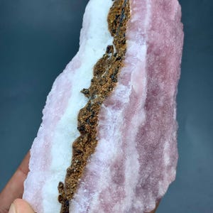 May include: A raw, natural mineral specimen featuring layers of pink, white, and brown hues. The pink sections have a textured appearance, while the white areas are smoother. A dark brown vein runs through the center of the stone.